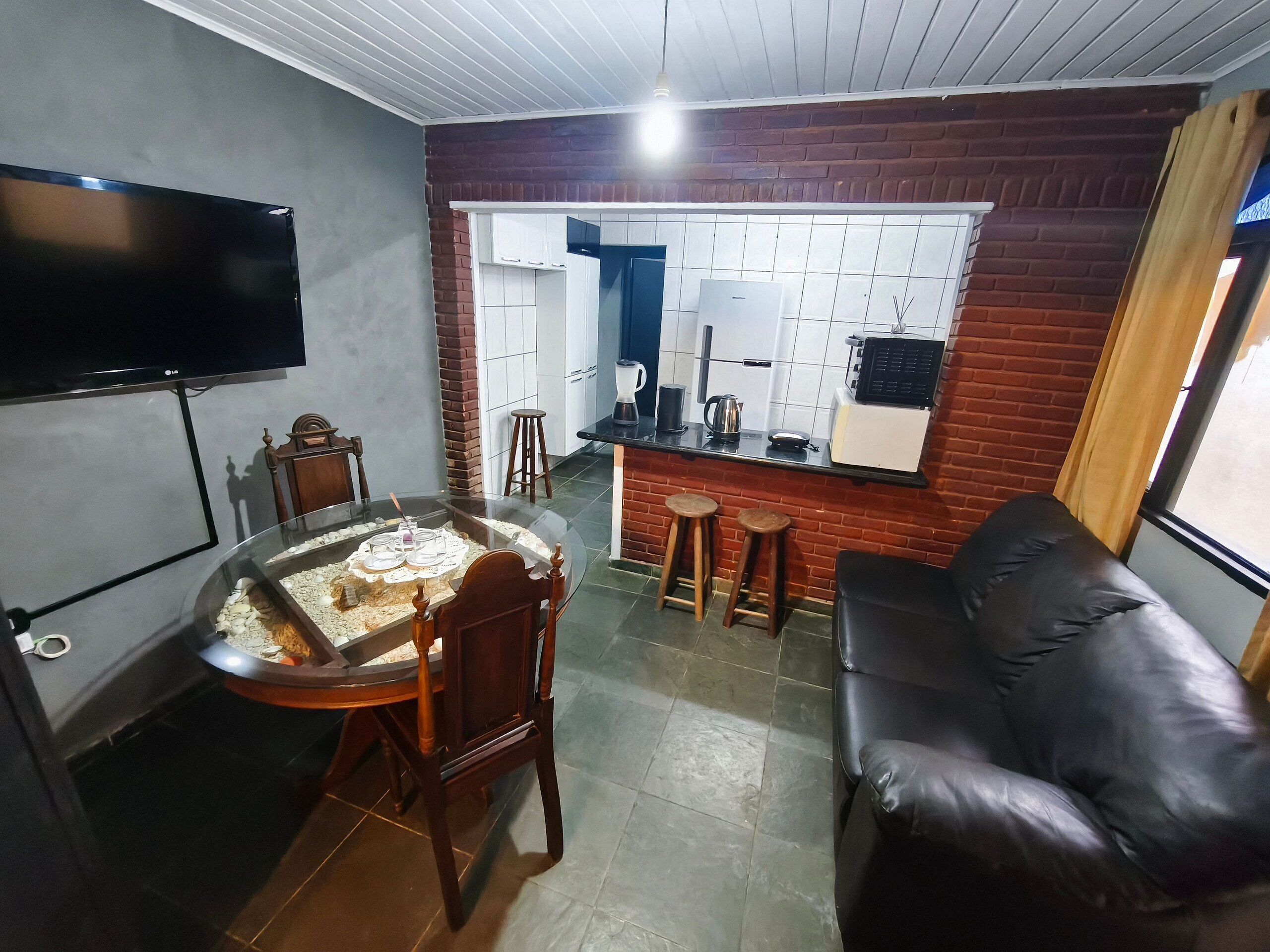 Een woonkamer met een eettafel, een zwarte leren bank, een aan de muur bevestigde televisie en een keuken met een bakstenen muur en betegelde achterwand.