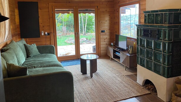 Interior acogedor de una cabaña con un sofá verde, una mesita redonda pequeña, un televisor sobre un soporte de madera y una chimenea con azulejos decorativos.