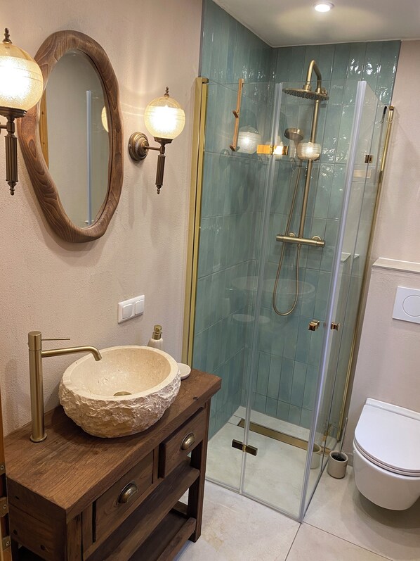 A bathroom with a stone sink, wooden vanity, two wall-mounted lights, a glass-enclosed shower, and a toilet.