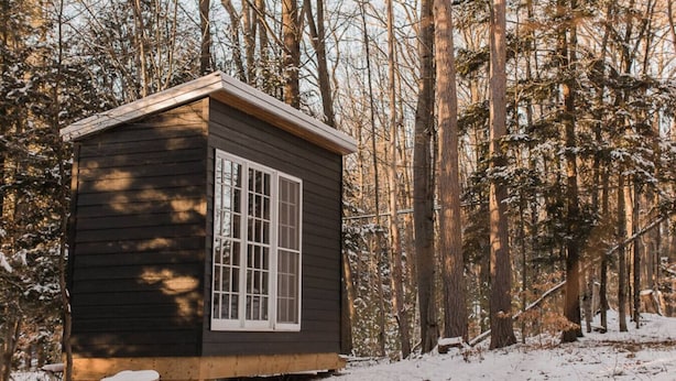 Una pequeña cabaña de madera con un ventanal grande, rodeada por un bosque cubierto de nieve.