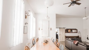 Un comedor amplio con una larga mesa de madera y sillas, un ventilador de techo y una puerta corrediza tipo granero que da a una sala de estar con un sofá y televisor.