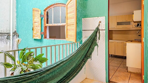 Un hamac dans une pièce dont un mur est peint en bleu sarcelle, une fenêtre avec des volets en bois, et un coin cuisine visible à travers une porte ouverte.