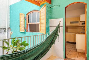 Una cadira lligadora en una habitació amb una paret color cel, una finestra amb persianes de fusta, i una cuina visible a través d'una porta oberta.