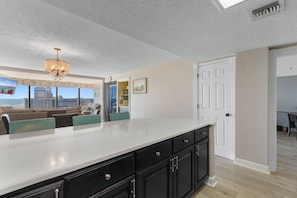 A modern kitchen with a large island, black cabinets, and a view of the beach. - Oceanfront Barefoot Serenity (Myrtle Beach)