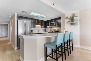 A modern kitchen with a central island, stainless steel appliances, and blue bar stools. - Oceanfront Barefoot Serenity (Myrtle Beach)