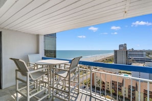 A balcony with a table and chairs, overlooking a beach and cityscape. - Oceanfront Barefoot Serenity (Myrtle Beach)