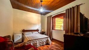 Quarto com teto de madeira, cama com colcha xadrez, mesa de cabeceira com abajur, janela com cortinas e televisão sobre um móvel.