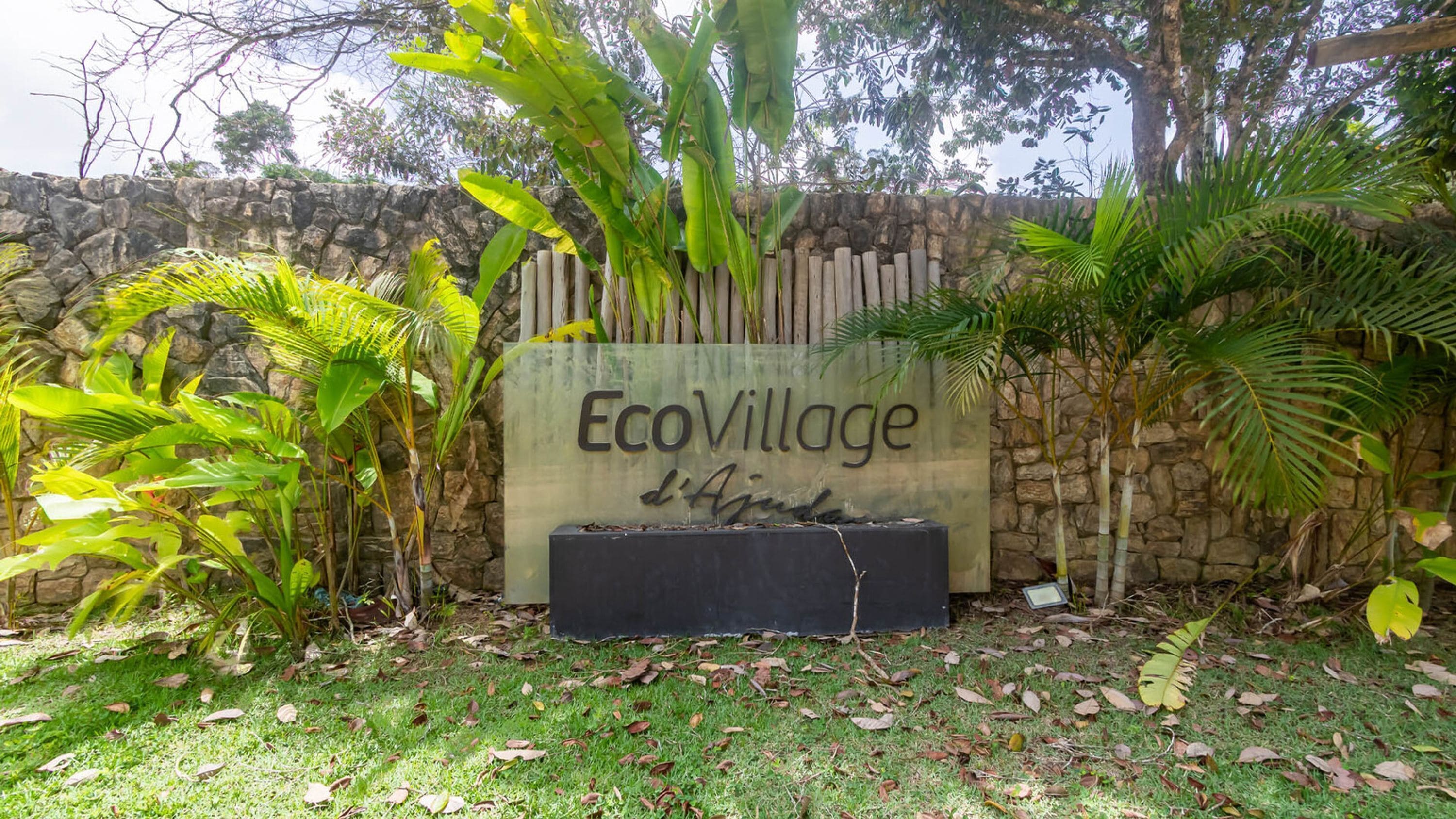 A sign for EcoVillage d'Ajuda with a stone wall and tropical plants around it.