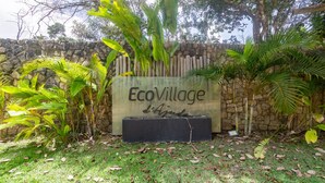 Señal de EcoVillage d'Ajuda, con un muro de piedra y plantas tropicales a su alrededor.