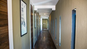 A long, narrow hallway with framed artwork on the walls, wooden paneling on one side, and a blue-tinted window on the other. - Loft com Smart Lock e Ar Perto do Projeto Tamar (Ubatuba)