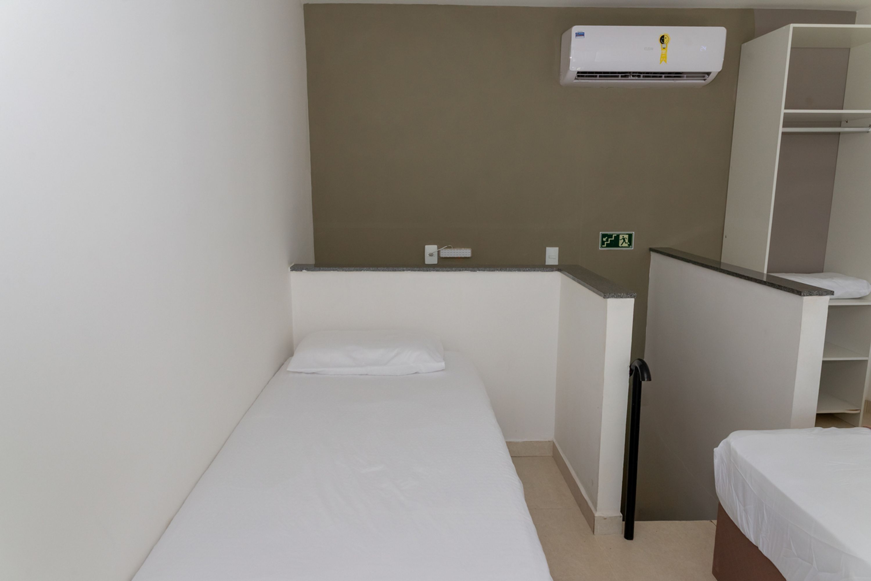 Een kleine kamer met een enkel bed, een witte muur en een witte airconditioning unit erboven.