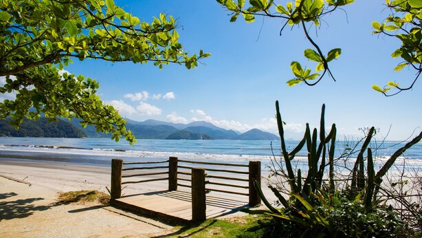 A beach with a wooden fence, a clear sky, and mountains in the background. - Loft com Smart Lock e Ar Perto do Projeto Tamar (Ubatuba)