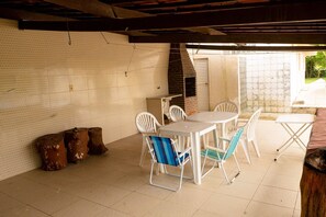 Un espace patio doté d’un sol carrelé, d’un four en briques, de meubles en plastique blancs et d’un mur blanc avec une fenêtre.