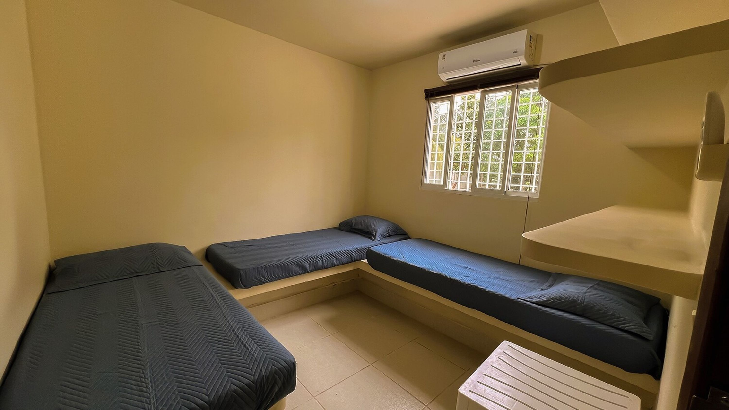 A small, single-room accommodation with two single beds, a window with a view of greenery, and an air conditioning unit above the window.