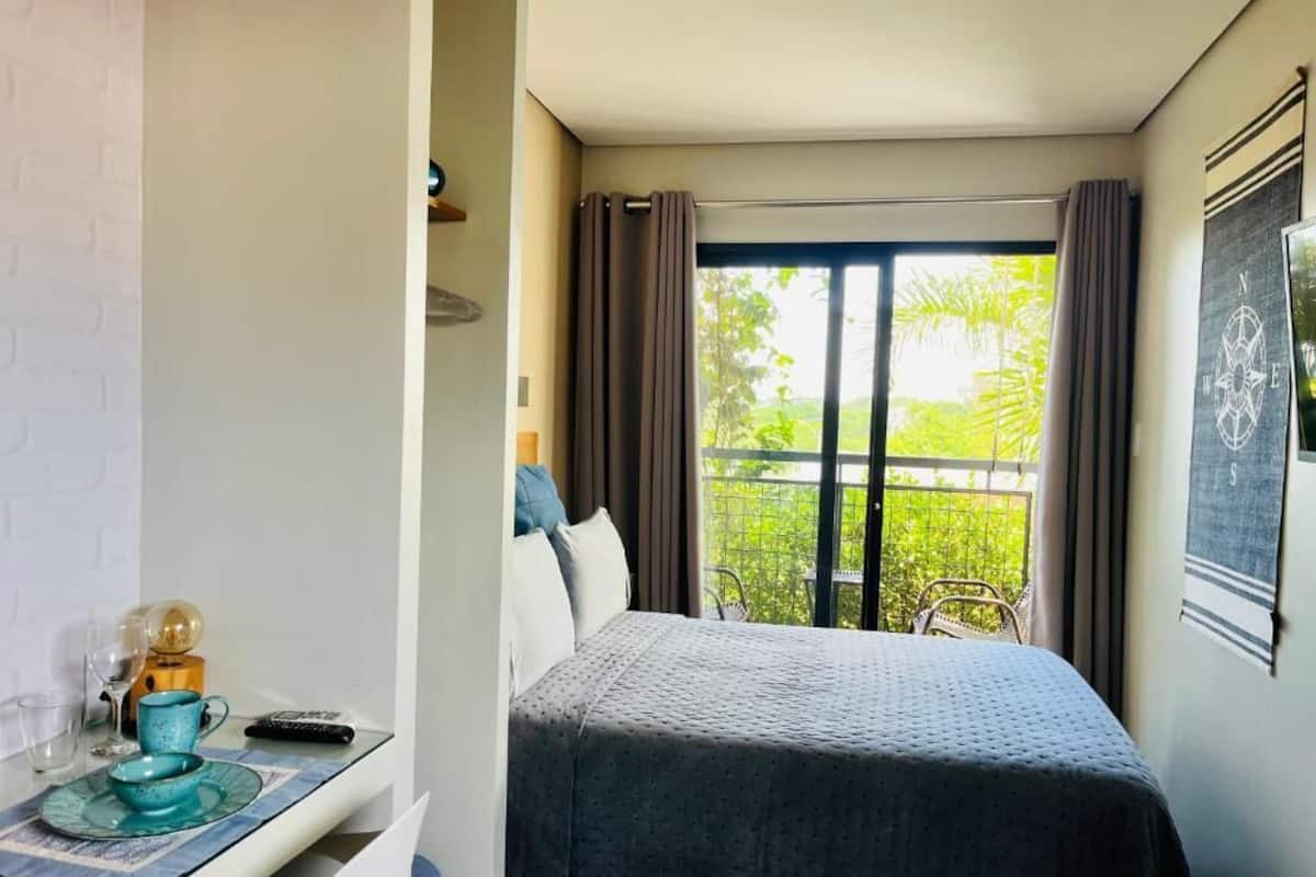 Una habitación pequeña y moderna con cama, una mesa y algunos objetos, y un ventanal con cortinas.