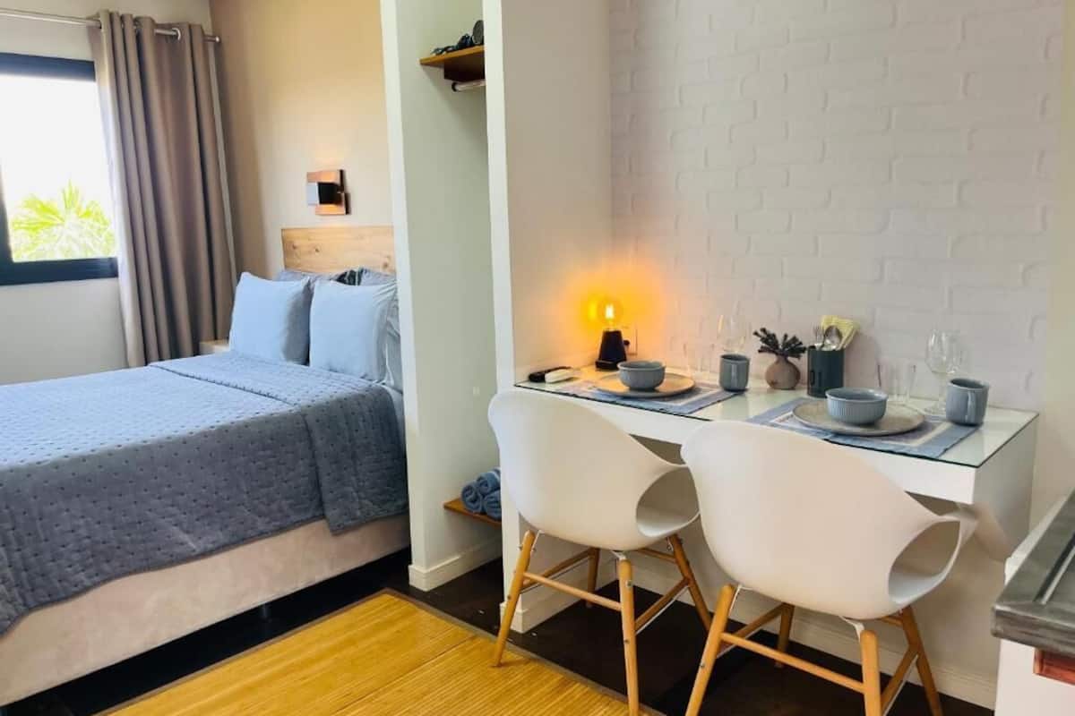 Un dormitorio con una cama, un escritorio con dos sillas, una lámpara y una pequeña estantería con objetos.