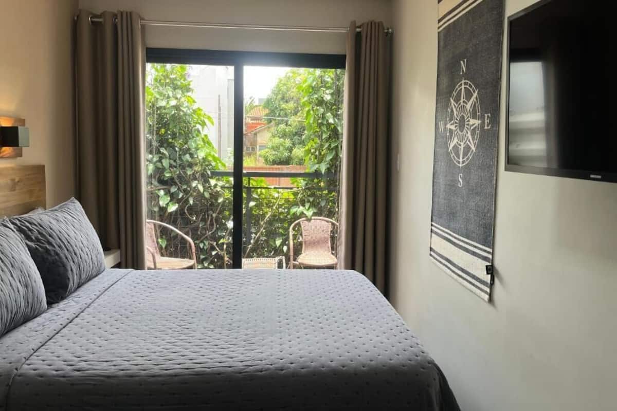 Una habitación de hotel con cama, televisión, ventana grande con cortinas y vista a zonas verdes.