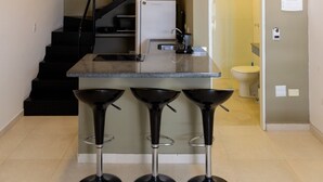 A modern kitchen with a central island, black bar stools, a staircase, and a visible bathroom. - Loft c Smart TV e Wi-fi pr x ao Aqu rio de Ubatuba (Ubatuba)