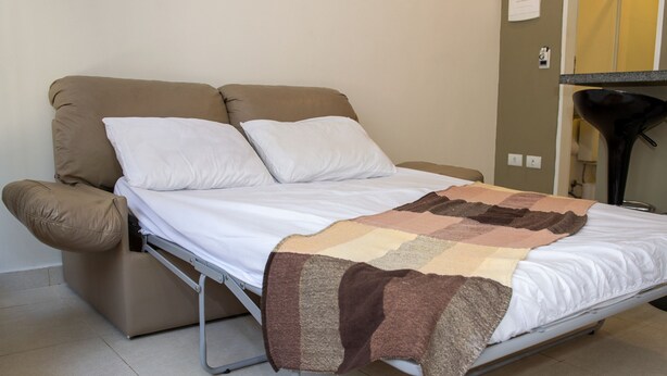 A single bed with a white mattress and a brown quilt, a beige sofa, and a small bedside table with a lamp. - Loft com Wi-fi e Smart Lock pr x ao Projeto Tamar (Ubatuba)