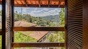 A view through a wooden-framed window with shutters, overlooking a landscape with terraced roofs, greenery, and hills. - Casa com Piscina Sal o de Jogos e Churrasqueira (Mairipora)