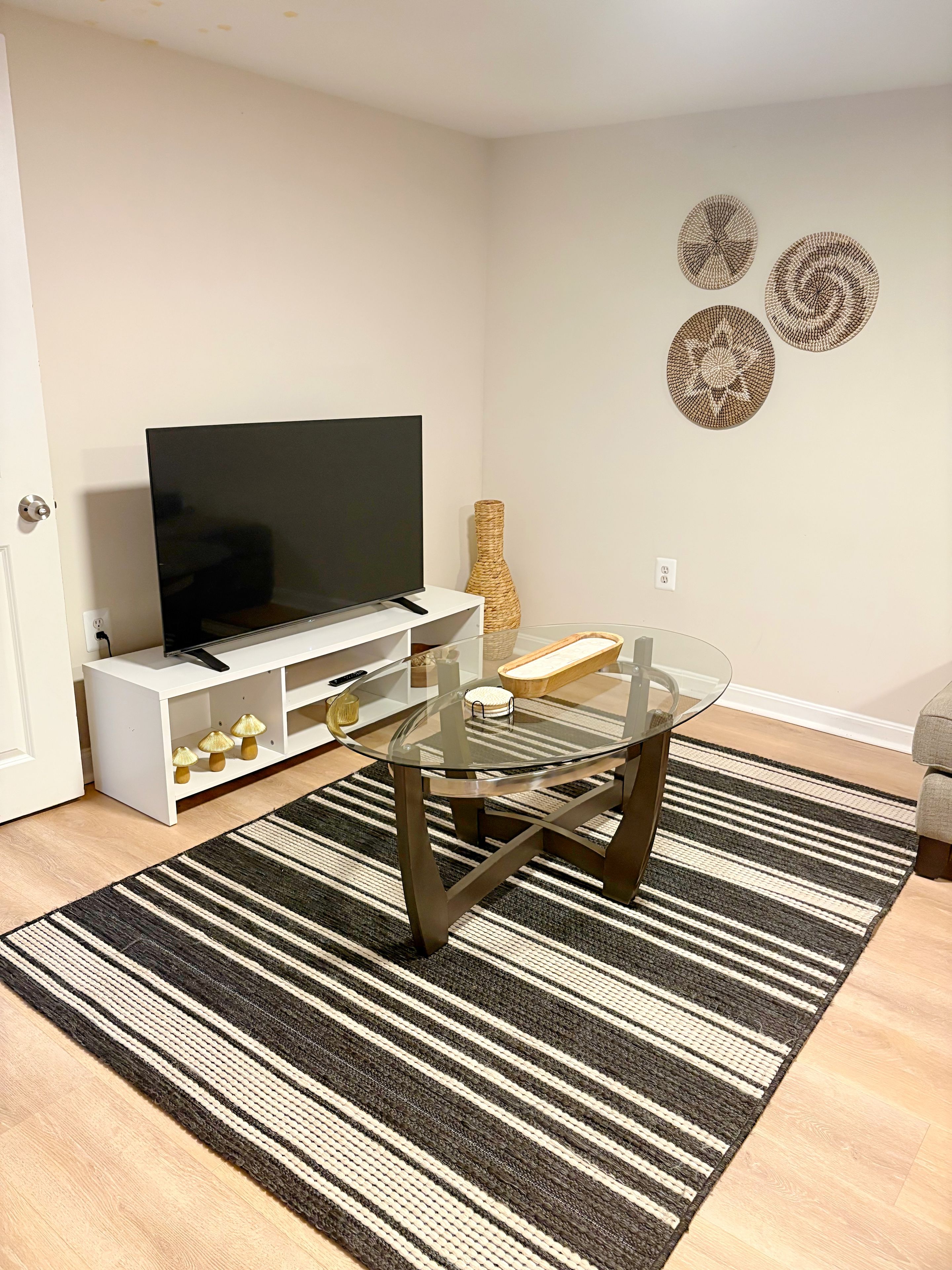 A modern living room with a glass-top coffee table, a flat-screen TV on a white console, a striped rug, and decorative wall art.