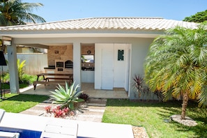 A house with a covered patio, a brick fireplace, and a small garden with palm trees and plants. - Casa com Piscina Quadra de Areia e Churrasqueira (Araruama)