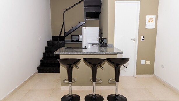 A kitchen area with a microwave, a coffee machine, and a refrigerator. There are three black bar stools with metal legs. A white door is visible in the background. - Loft com Wi-fi e Smart Lock Perto do Projeto Tamar (Ubatuba)