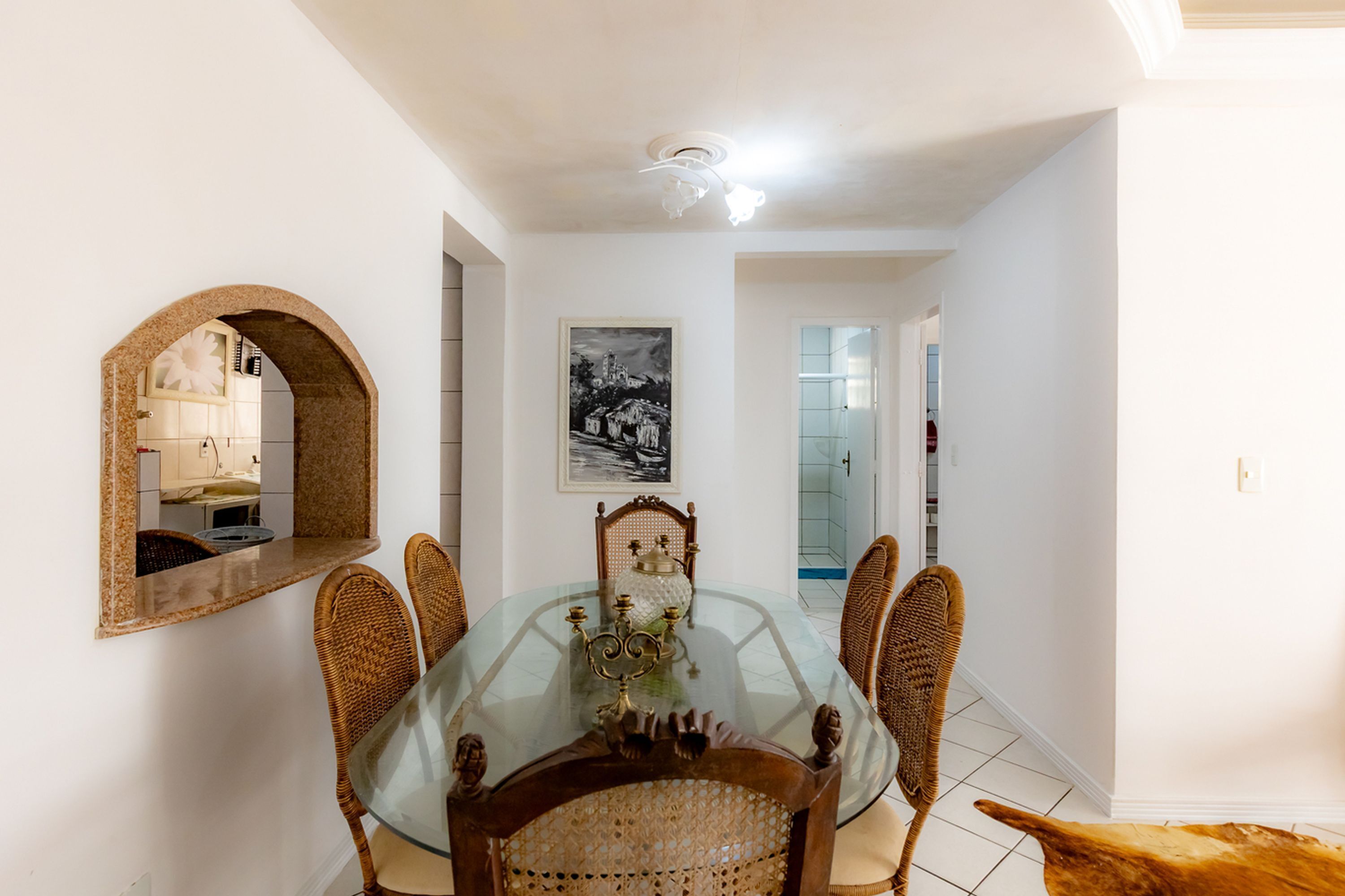 Un'area pranzo con un tavolo in vetro circondato da sedie in vimini, uno specchio decorativo e un quadro appeso al muro.