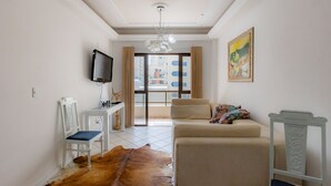 A living room with a sofa, a television, a desk, and a chair. - Apartamento com Varanda Churrasqueira e Vista Mar (Itapema)