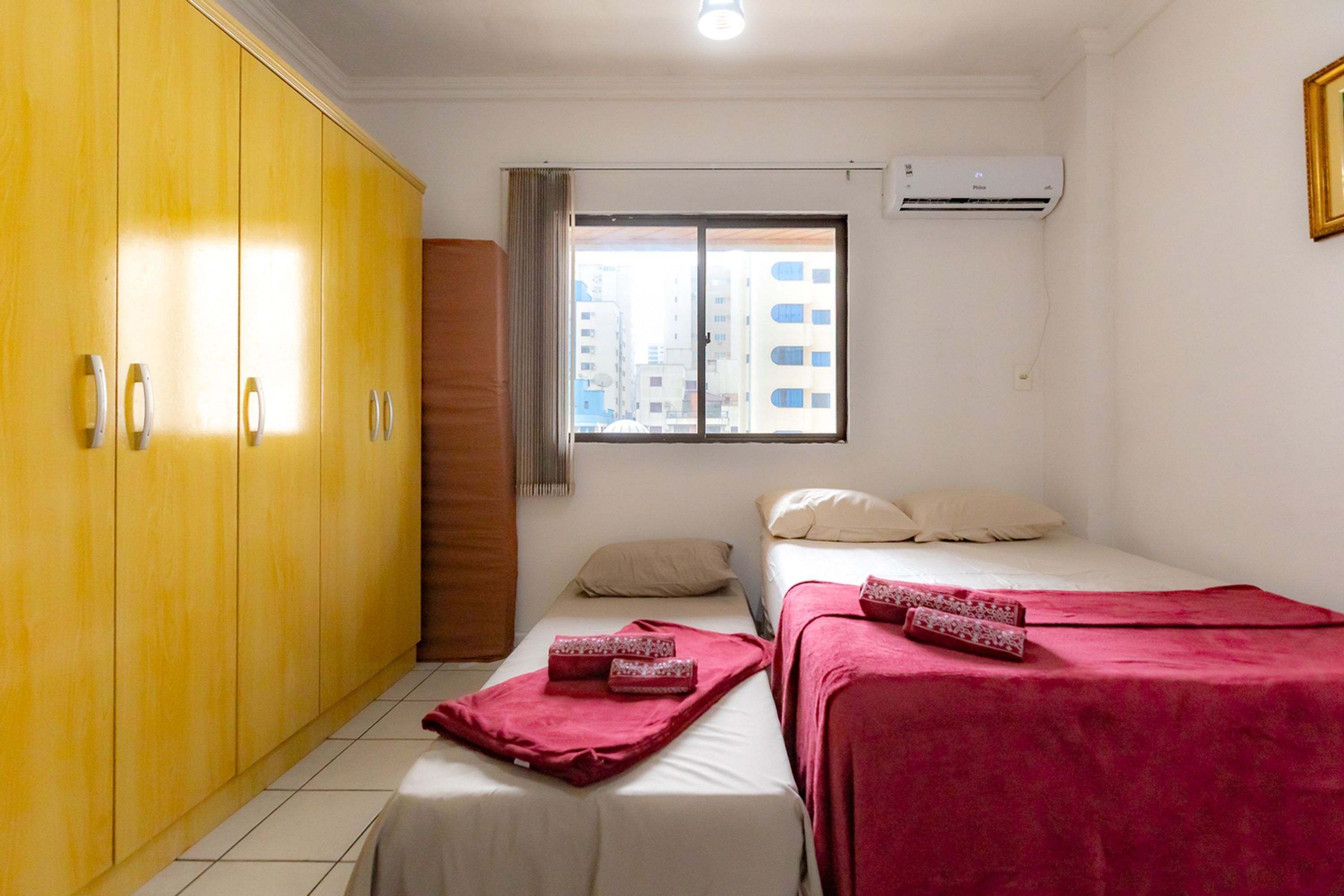 A hotel room with two beds, a wardrobe, a window with a view of buildings, and an air conditioning unit.