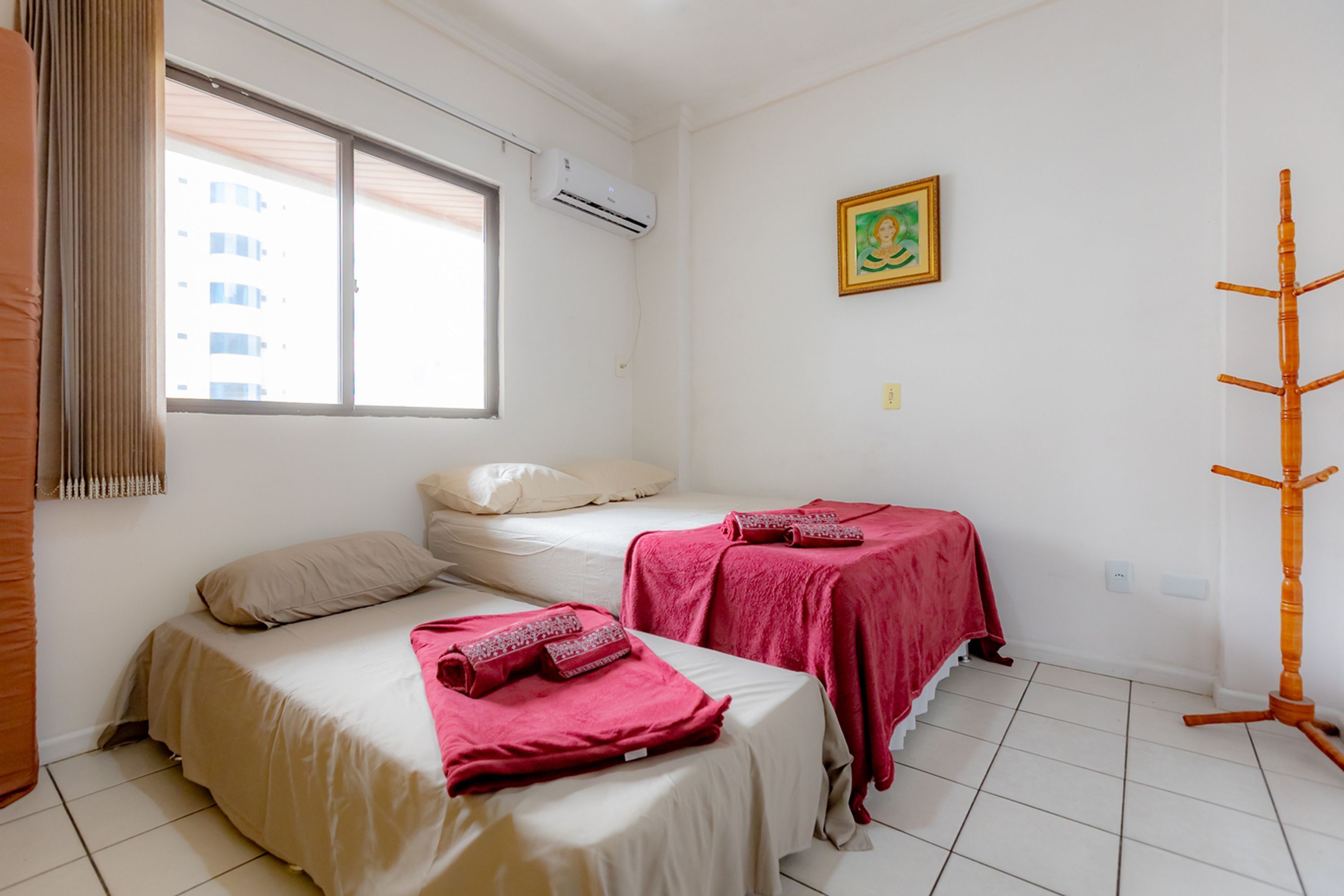 A hotel room with two beds, one with a beige bedspread and the other with a red bedspread, a window with blinds, an air conditioning unit, a wall-mounted picture, and a wooden rack.