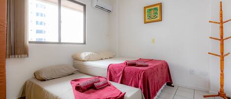 A hotel room with two beds, one with a beige bedspread and the other with a red bedspread, a window with blinds, an air conditioning unit, a wall-mounted picture, and a wooden rack.