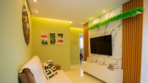 A modern living room with a sofa, a TV mounted on a marble wall, and a decorative plant. - Apto com Piscina e Garagem em Barra dos Coqueiros (Barra dos Coqueiros)