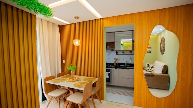 A modern dining area with a wooden table and chairs, a small plant, and a kitchen visible through an open doorway. - Apto com Piscina e Garagem em Barra dos Coqueiros (Barra dos Coqueiros)