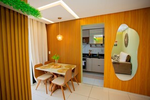 Uma sala de jantar moderna com uma mesa e cadeiras de madeira, uma pequena planta e uma cozinha visível através de uma porta aberta.