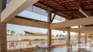 An outdoor cafe area with tables and chairs, a pool in the background, and a view of palm trees and buildings. - Apto com Piscina e Garagem em Barra dos Coqueiros (Barra dos Coqueiros)