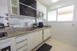 A modern kitchen with white cabinets, a granite countertop, a built-in microwave, and a window providing natural light. - Apto com Varanda e Churrasqueira a 100m da Orla (Praia Grande)