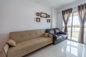 A living room with a beige sofa, a dark leather sofa, and a large window with curtains. - Apto com Varanda e Churrasqueira a 100m da Orla (Praia Grande)