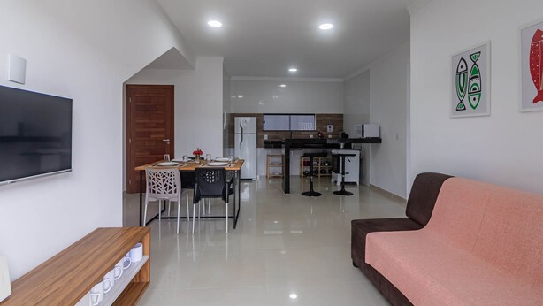A modern living room with a flat-screen TV, a brown sofa, and a dining area with a wooden table and chairs. - Pedra DO MAR Apto Perto da Praia de Taperapuan (Porto Seguro)
