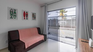 A modern living room with a dark brown sofa, a pink throw blanket, and a flat-screen TV on a wooden table. There are three framed artworks on the wall and a sliding glass door leading to an outdoor area with a metal gate and palm trees. - Pedra DO MAR Apto Perto da Praia de Taperapuan (Porto Seguro)