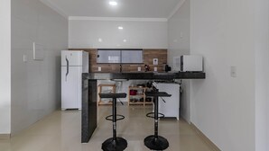 A modern kitchen with a refrigerator, microwave, and a wooden backsplash. There are two black bar stools and a small wooden table. - Pedra DO MAR Apto Perto da Praia de Taperapuan (Porto Seguro)