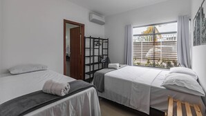 A hotel room with two beds, a wooden bench, a window with blinds, and a door leading to another room. - Pedra DO MAR Apto Perto da Praia de Taperapuan (Porto Seguro)
