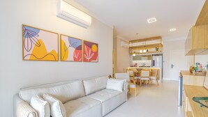 Ein modernes Wohnzimmer mit grauem Sofa, einem Essbereich mit Holztisch und -stühlen sowie einer Küche mit eingebautem Kühlschrank und Holzschränken.