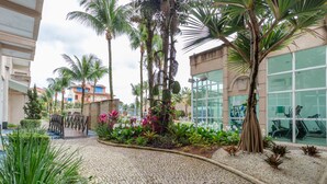 A paved courtyard with palm trees, a brick pathway, and a glass building in the background. - Loft c Varanda Piscina e Academia em Itacuru (Mangaratiba)