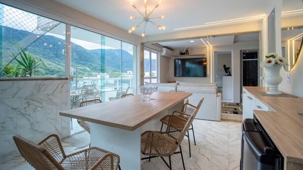 A modern kitchen with a large island, a dining area with wicker chairs, and a view of mountains through large windows. - Loft c Varanda Piscina e Academia em Itacuru (Mangaratiba)