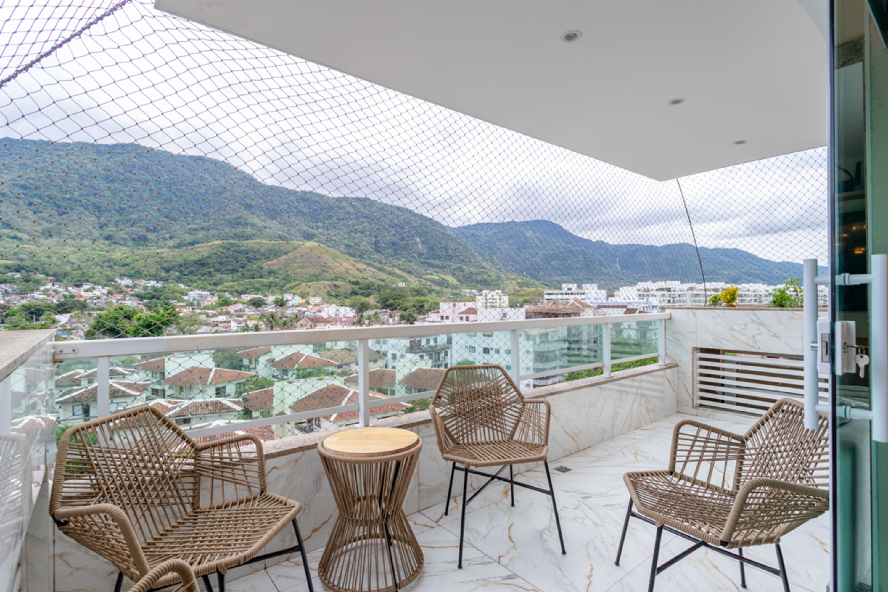 Varanda com cadeiras de vime e mesa redonda, com vista para a paisagem urbana e montanhas.