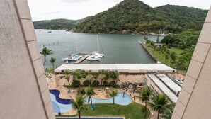 A view of a hotel with a swimming pool, a dock with boats, and a mountainous landscape in the background. - Loft c Varanda Piscina e Academia em Itacuru (Mangaratiba)