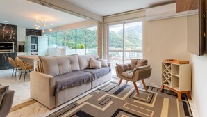 A modern living room with a sofa, armchairs, and a coffee table. There is a large window with a view of mountains. - Loft c Varanda Piscina e Academia em Itacuru (Mangaratiba)