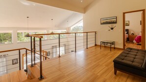 A spacious loft with high ceilings, wooden flooring, a black leather sofa, a glass table, and a view of greenery outside.