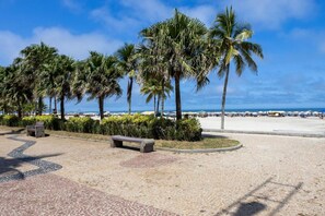 A paved walkway lined with palm trees and benches, leading to a beach with umbrellas and people in the distance. - Apto com Ar-condicionado e Churrasqueira Privativa (Praia Grande)